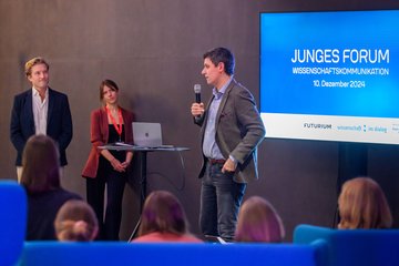 Drei Personen, eine mit Mikrophon in der Hand. Auf einem Display im Hintergrund die Schrift "Junges Forum Wissenschaftskommunikation, 10. Dezember 2024"