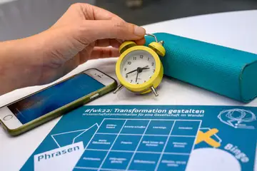 Bingo-Flyer, Stoppuhr und ein Smartphone auf einem Tisch. Eine Hand hält die Stoppuhr.