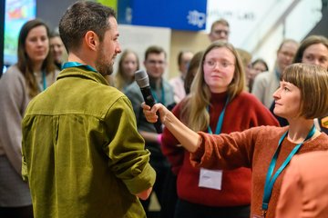 Zwei Personen beim Interview, weitere Personen im Hintergrund.