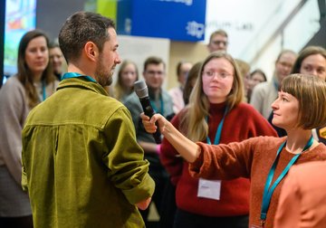 Zwei Personen beim Interview, weitere Personen im Hintergrund.