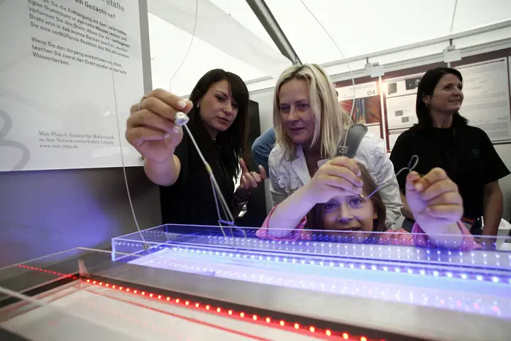 Mehrere Frauen und ein Kind bei einer Wissenschaftssausstellung in einem Zelt. Eine Frau zeigt ein Experiment.