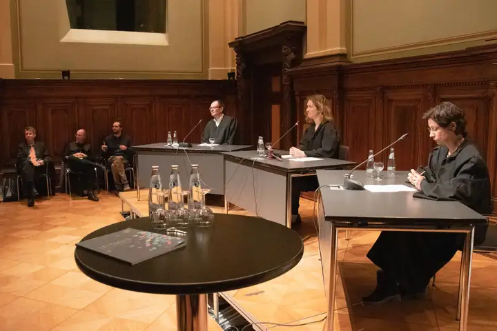 Eine fiktive Gerichtsverhandlung: Drei Personen in dunklen Roben sitzen an drei Tischen.