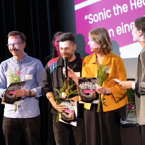 Mehre Personen lachen auf Bühne und nehmen Blumen und Auszeichnung entgegen