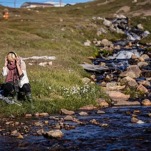 Eine Frau sitzt in der Natur neben einem Bach und hört über Kopfhörer etwas an.