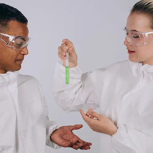 Zwei Wissenschaftler*innen in Kittel und mit Schutzbrille. Eine Person hält ein Reagenzglas mit einer grünen Flüssigkeit in der Hand.