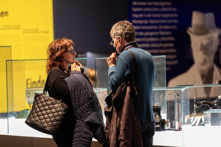 Zwei Personen, die sich gegenüber stehen und über Kopfhörer etwas anhören. Im Hintergrund sind Vitrinen des Industriemuseums zu sehen.