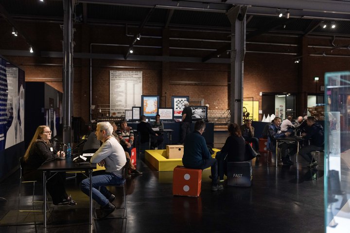 Der Ausstellungsraum des Industriemuseums. Mehrere Menschen sitzen in Kleingruppen verteilt im Raum. Einige Personen unterhalten sich an Tischen, andere Sitzen mit Kopfhörern auf Sitzwürfeln.