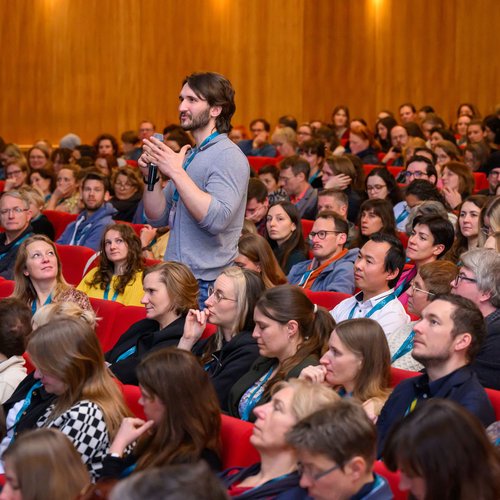 Das Publikum in einem großen Saal. Ein Mann ist aufgestanden und spricht in ein Mikro.
