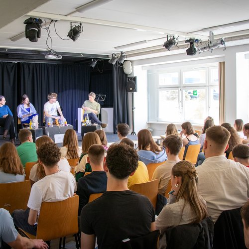 Schüler*innen und Forscher*innen im Gespräch auf Bühne vor Schüler*innen in Klassenraum