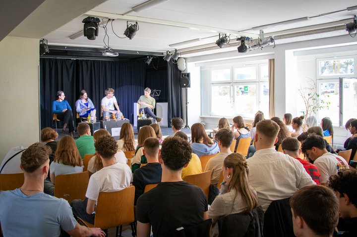 Schüler*innen und Forscher*innen im Gespräch auf Bühne vor Schüler*innen in Klassenraum