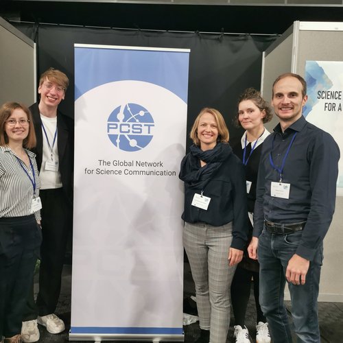 Fünf Personen, Liliann Fischer, Vincent Schmid-Loertzer, Anne-Sophie Behm-Bahtat, Julia Panzer und Bastian Kremer, stehen vor einem Banner mit der Aufschrift PCST The Global Network for Science Communication