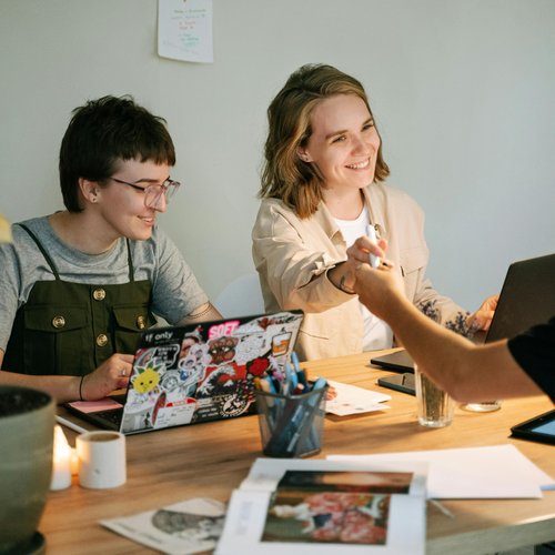 Drei junge Menschen sitzen an einem Tisch mit Laptops und arbeiten gemeinsam; zwei von ihnen geben sich lächelnd die Hand.