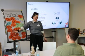 Julia Panzer, stellvertretende Projektleitung der Impact Unit steht vor einem Präsentationsbildschirm und einer Flipchart. Mit ausgestrektem Arm zeigt sie auf die Flipchat. Zur Frage "Welche Erfahrungen habt ihr mit Evaluation?" sind dort viele runde und
