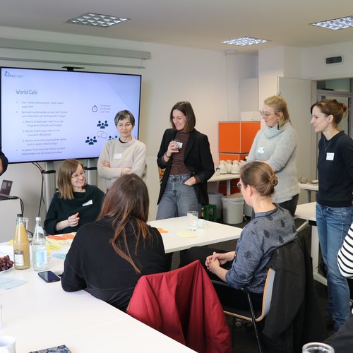 Eine Gruppe aus 9 Personen versammelt sich um einen Tisch, auf dem eine Mind-Map aus bunten runden Zetteln trapiert ist. Drei Personen sitzen am Tisch, der Rest der Gruppe steht um sie herum.