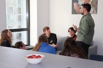 Vincent Schmid-Loertzer sitzt aufmerksam mit einer sechsköpfigen Gruppe zusammen. Eine Person aus der Gruppe steht und spricht mit Gestiken in die Runde.