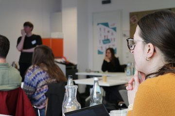 Im Vordergrund, rechts unten im Bild ist eine Teilnehmerin, die konzentriert einem Vortrag lauscht. Unscharf im Hintergrund sind weitere Personen in der Rückenansicht gezeigt.