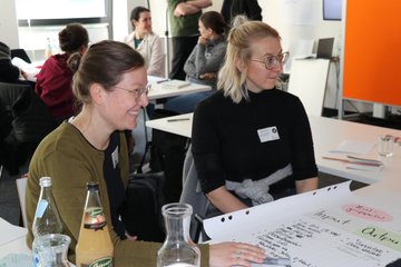 Zwei Teilnehmerinnen mit Brille sind im Profil gezeigt. Sie sitze um ein Plakat. Im Hintergrund sieht man eine andere Gruppe aus Teilnehmenden, die sich miteinander besprechen.