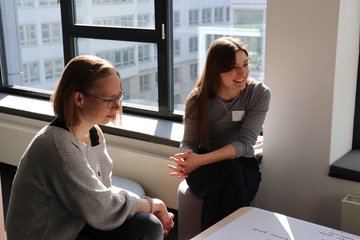 Zwei Teilnehmerinnen sitzen schräg vor einer Fensterfront vor einem Tisch mit Plakat. Sie scheinen sich gerade mit Personen außerhalb des Bildes zu unterhalten.
