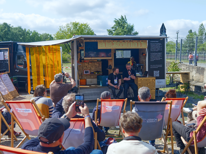 Foto des Energiemobils, zwei Personen mit einem Mikro sitzen davor. Im Vordergrund Menschen auf Liegestühlen.