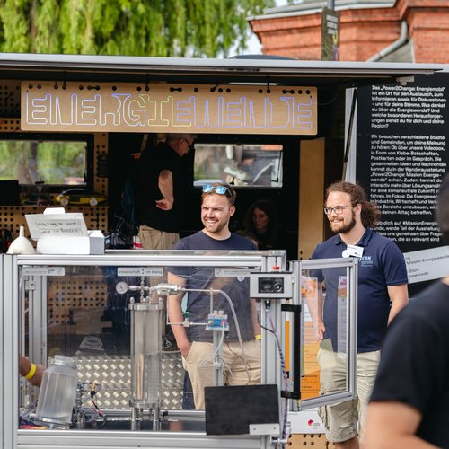 Personen stehen mit Glaskasten mit Präparaten für Wasserstoffgewinnung vor Anhänger, der als fahrende Ausstellung fungiert.