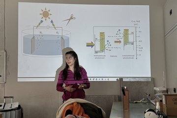 Eine weiblich gelesene Person mit langen dunklen, halboffenen Haaren steht vor einer Beamerleinwand und wird entsprechend vom Beamer angestrahlt. Um sie herum wird eine Folie angezeigt, die ein chemisches Erklärungsmodell zeigt.