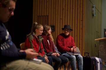 Drei Personen, sitzend. Sie alle tragen rot, hinter ihnen ist eine Holzwand. Die männlich gelesene Person rechts im Bild trägt zu dem roten Pulli einen schwarzen Hut, verschränkt die Hände auf dem Schoß und scheint den anderen beiden Personen im Bild gera