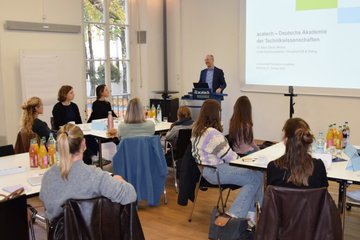 Personen an Tischen in Seminarraum, Vortragender vor Präsentation