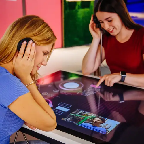 Zwei Personen in einer Ausstellung: Die Personen stehen an einem großen Touchscreen und haben Kopfhörer an.