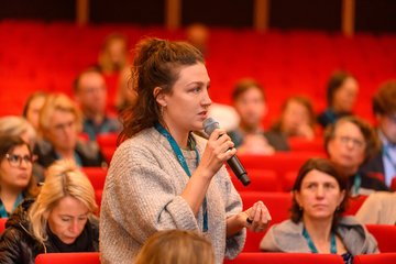 Publikum in Saal sitzend, Frau stehend mit Mikrofon in der Hand