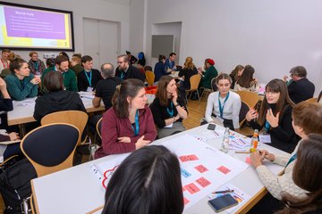 Personen um einen Tisch sitzend und stehend, auf dem ein Plakat mit bunten Post-It Zetteln liegt