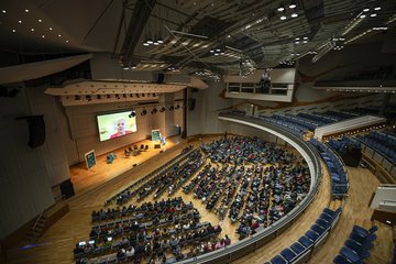 Gefüllter Hörsaal und Person wird auf Folien der Präsentation angezeigt
