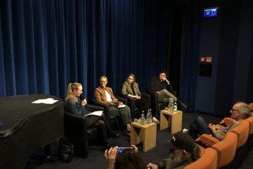 Auf einer Bühne, die von blauen Vorhängen umrahmt wird, sitzen vier Personen nebeneinander auf schwarzen Sesseln mit je einem Mikrofon in der Hand. Die Person links im Bild spricht als einzige in ihr Mikrofon rein. Ein Publikum ist leicht angeschnitten.