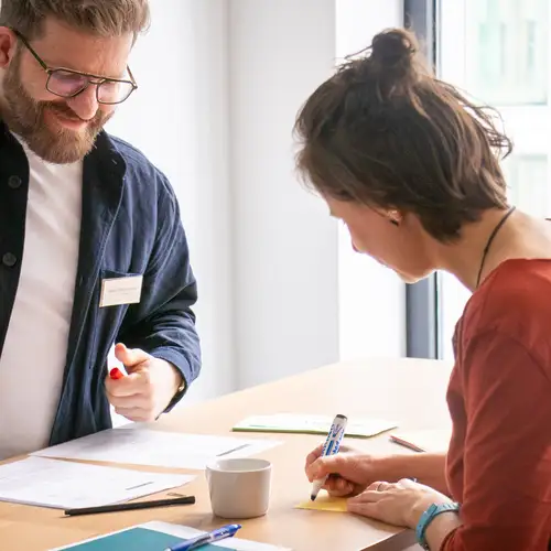 Ein Mann und eine Frau stehen sich an einem Tisch gegenüber und notieren etwas auf Zetteln.