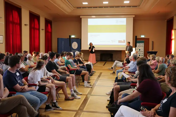 Personen sitzen sich in einem großen Saal auf zwei Seiten gegenüber. Vorne stehen zwei Moderator*innen.