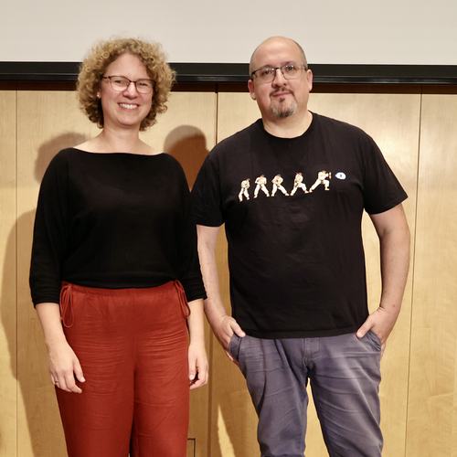 Dr. Jana Heysel und Dr. Sascha Vogel posieren gemeinsam für ein Foto. Jana trägt kurze blonde Locken und eine Brille. Sascha trägt eine Glatze, Brille und hält die Hände in den Hosentaschen.