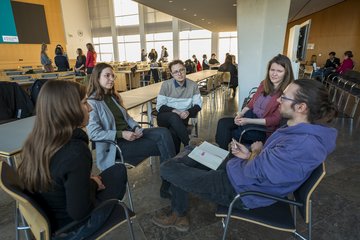 Im Vordergrund ist ein Sitzkreis mit fünf Personen, die sich angeregt unterhalten. Im Hintergrund ist ein weiter Sitzungssaal zu sehen.
