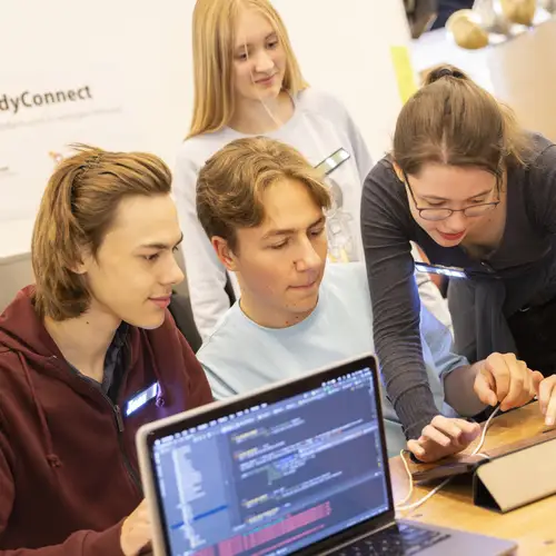 Mehrere Jugendliche tüfteln gemeinsam an einem Tablet. Im Vordergrund ist ein Bildschirm zu sehen, der Programmier-Codes zeigt.