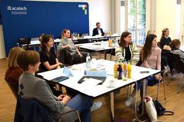 Personengruppe sitzend um Tische in Seminarraum, schauen alle in eine Richtung