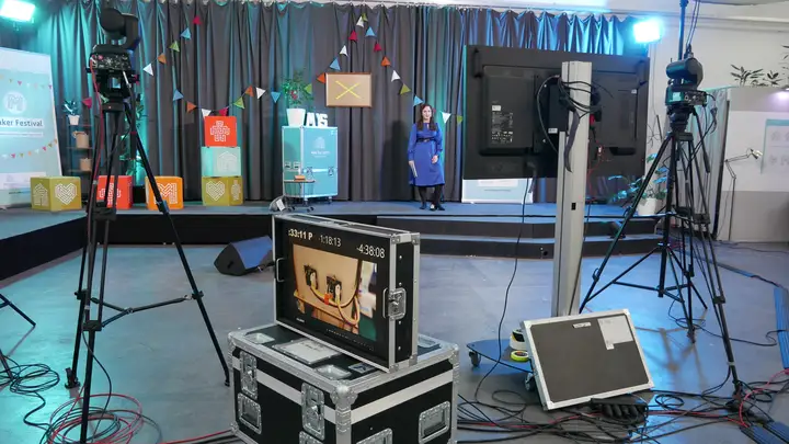 Eine Moderatorin auf einer bunt geschmückten Bühne. Vor der Bühne sind mehrere Kameras und ein großer Bildschirm aufgebaut.