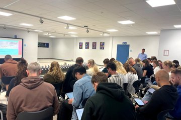 In einem Saal sieht man ein Sitzpublikum von hinten, einige haben aufgeklappte Laptops auf ihrem Schoß.