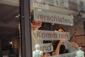 Ein Schaufenster, an dem gerade folgender Schriftzug von zwei Personen angebracht wird: Die Energiewende hat verschlafen. Komm rein und weck sie auf!