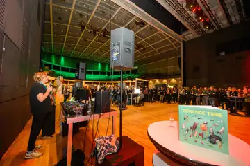 Ein Mann steht in einem großen Raum vorne an einem DJ-Pult. Gegenüber steht ein Publikum.