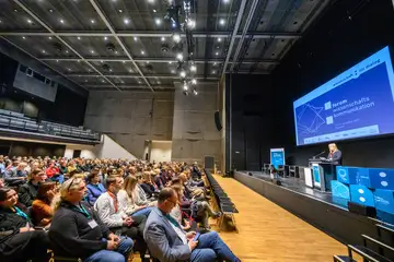 Ein voll besetzter Saal. Auf der Bühne steht eine Frau und spricht.