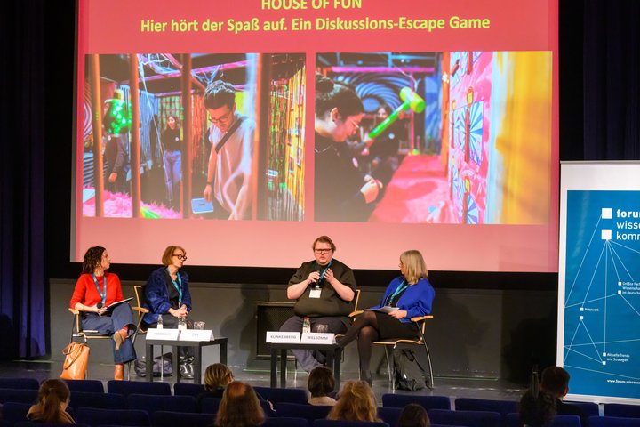 Vier Personen sitzen auf einer Bühne und diskutieren. Im Hintergrund ist eine Präsentation zu sehen. Sie zeigt zwei Fotos von Personen an einem Exponat, darüber steht "House of Fun. Hier hört der Spaß auf. Ein Diskussions-Escape Game."
