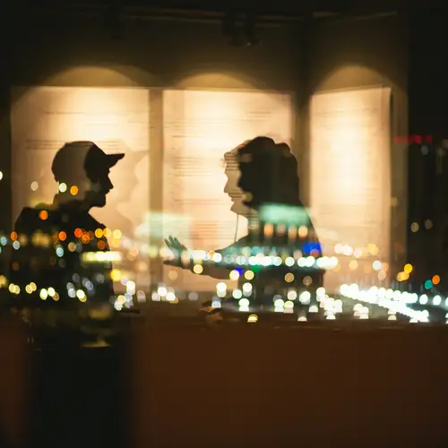 Eine Spiegelung im Fenster, die zwei Personen im Gespräch zeigt. Draußen sind Lichter der Stadt zu sehen.
